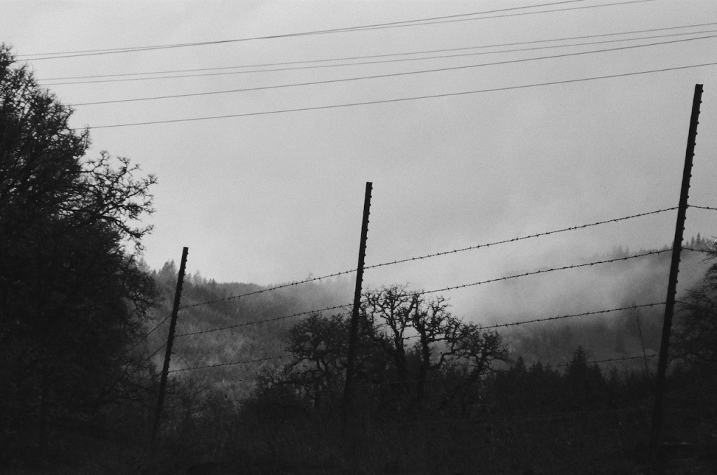 black and white image of barbed wire fence on a foggy day. a hill in the background is only partially visible.