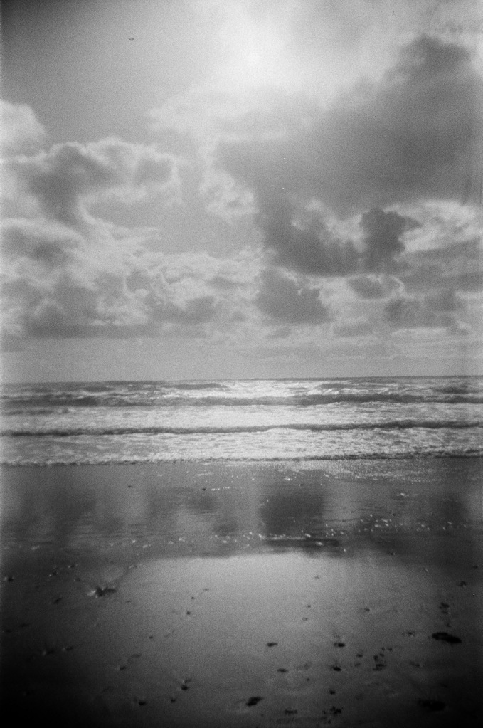 black and white image of a beach on a day with bright sun and scattered clouds. the sand is reflective, giving the image a dream-like quality